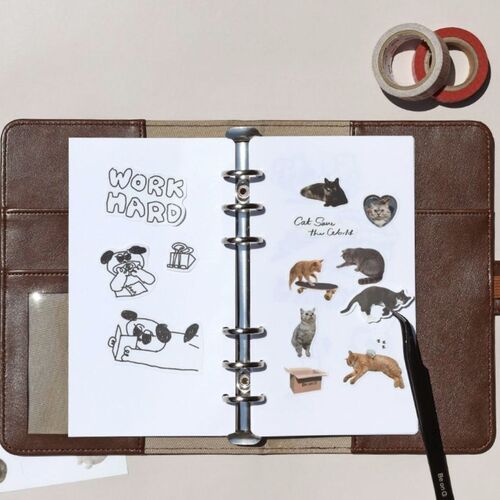 An open leather-bound binder displays a decorated page with playful cat and dog stickers, surrounded by washi tapes and a black pen.