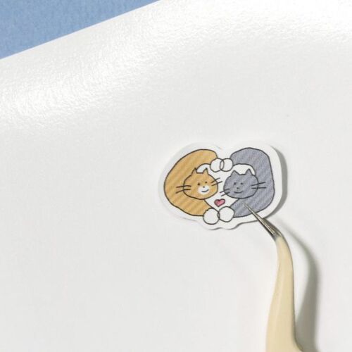 A close-up of a sticker featuring two adorable cats, one orange and one gray, decorated with a heart, on a white background.