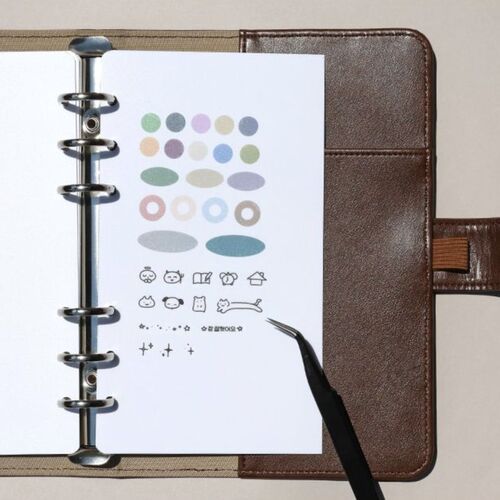 A white sticker page with colorful oval and icon designs rests inside a brown leather binder, illuminated by soft, even lighting.