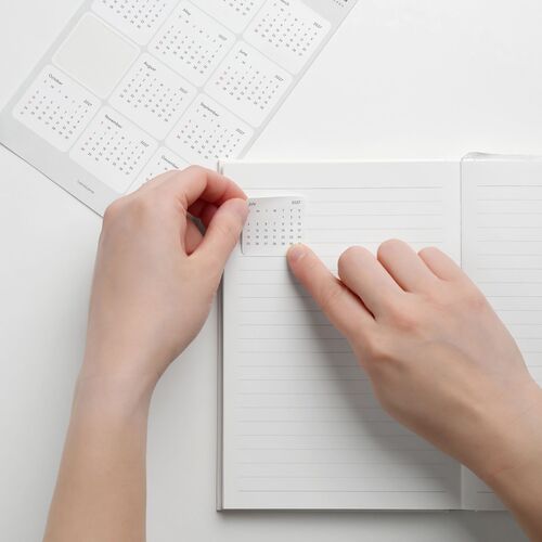 Hands place July 2027 calendar sticker onto a white lined planner page, emphasizing ease of customization and clarity.