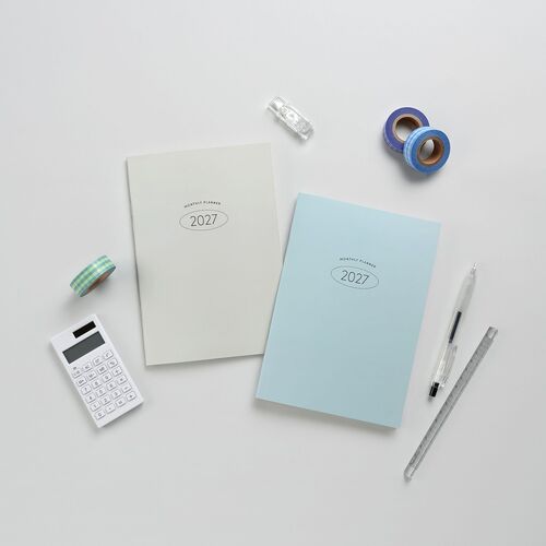 Two pastel-colored 2027 planners with minimalistic covers are displayed alongside stationery accessories on a clean, white surface.