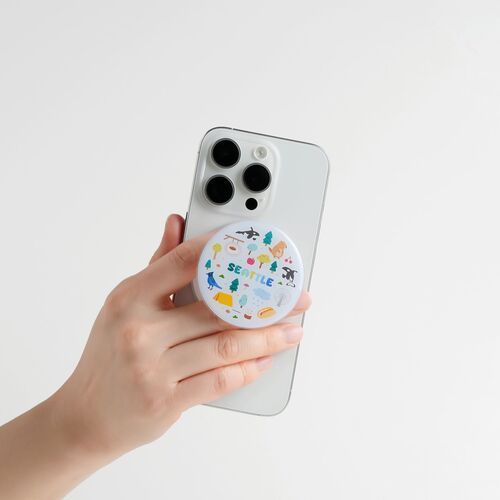 A hand holds a sleek white iPhone with a colorful Seattle-themed PopSocket attached, set against a plain white background.