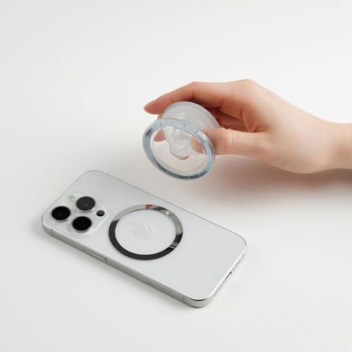 A hand holds a transparent, collapsible magnetic grip with a glossy epoxy finish, positioned above a sleek, white smartphone with triple camera lenses.