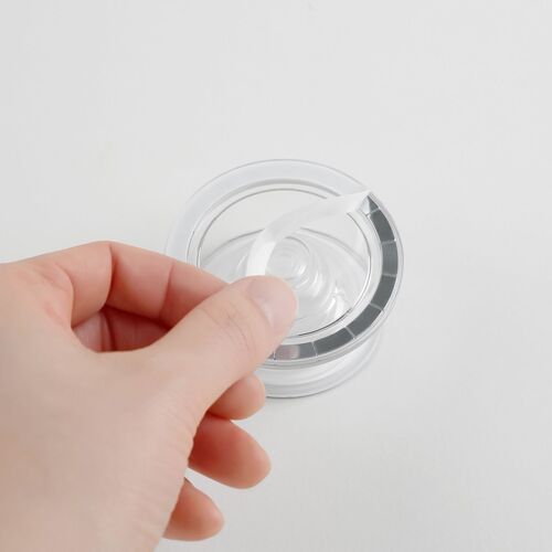 A hand peeling off a transparent adhesive backing from a clear, round magnetic phone grip with a white and silver rim, set against a plain white background.