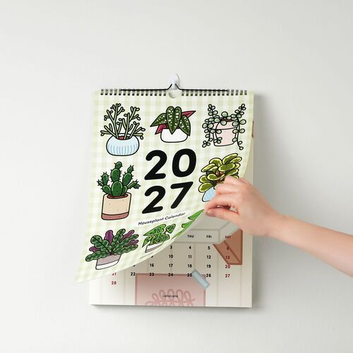 The image showcases a person flipping through a colorful, illustrated 2027 houseplant-themed wall calendar hanging on a plain white wall.