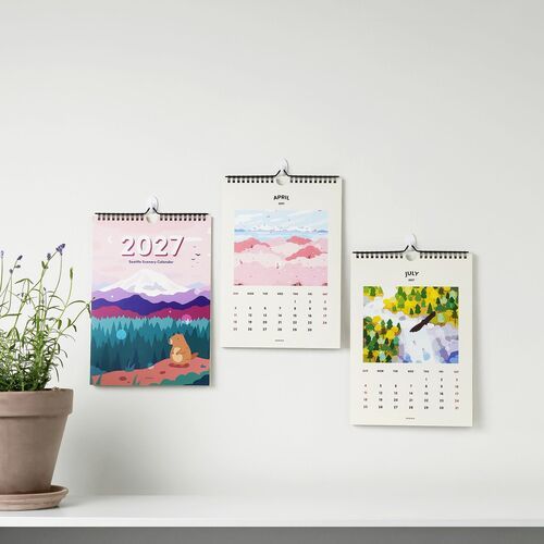 Colorful, artistic calendar pages with vibrant illustrations of Seattle scenes, hung neatly on a white wall with a potted plant nearby.