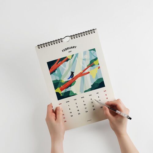 A person holds the February page of the 2027 Seattle Wall Calendar, featuring vibrant artwork of a stylized forest scene with a bird, against a plain white background.