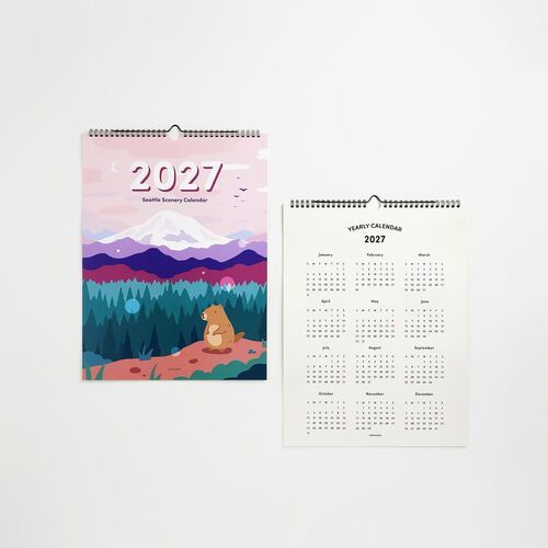 Two Seattle-themed wall calendars hang side by side, one featuring a colorful illustrated mountain scene with a bear, the other displaying a minimalistic 2027 yearly overview.