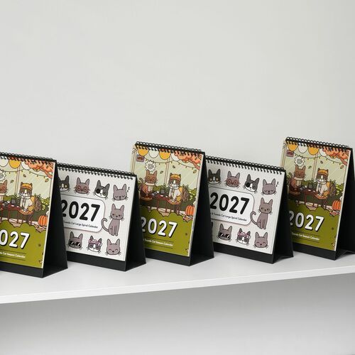 Multiple small desk calendars featuring colorful Lana and Tuxedo cats in playful scenes, arranged in a neat row on a white surface.