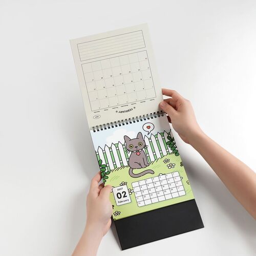 The image showcases a person flipping through a colorful, illustrated desk calendar featuring Lana and Tuxedo cats, with a vibrant outdoor scene and February's page visible.