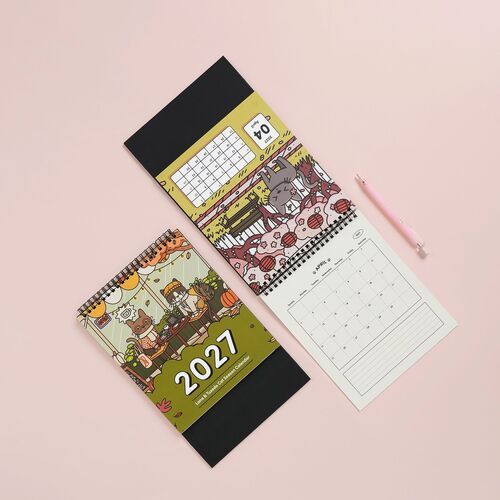 Two spiral-bound calendars with vibrant, playful cat-themed illustrations are displayed on a soft pink background, accompanied by a pink pen.