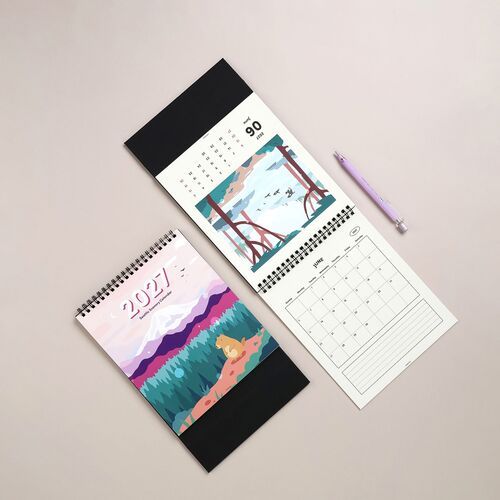 Two spiral-bound calendars displayed on a neutral surface, showcasing vibrant Seattle scenery and a colorful mountain landscape, with a purple pen nearby.
