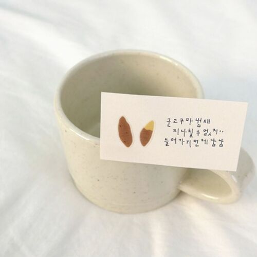 A small, off-white ceramic mug with a speckled finish holds a handwritten note featuring simple illustrations, set against a soft, white background.