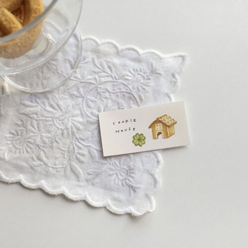 A delicate white embroidered cloth with scalloped edges holds a small, colorful note card labeled 'Cookie House,' accompanied by a glass of cookies on a white surface.
