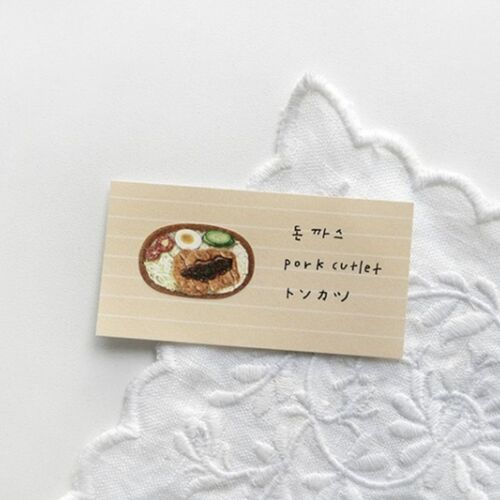 A small beige sticky note featuring a colorful illustration of a pork cutlet bowl, placed on a white embroidered fabric background.