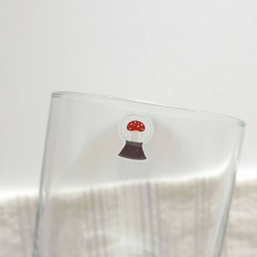 A cute sticker featuring a tiny, whimsical mushroom with red and white details, placed on the rim of a transparent glass.
