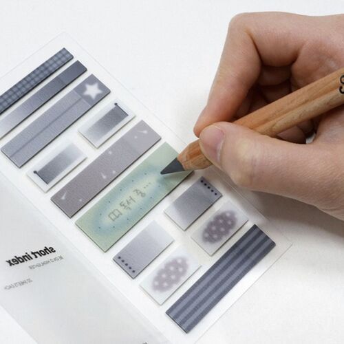 A hand writes on a light-colored sticky note with a wooden pencil, surrounded by various gray and patterned sticky notes on a white background.