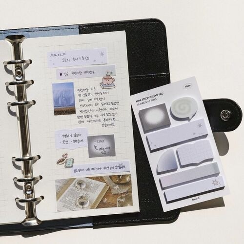 The image showcases a black, textured binder open to reveal a page with handwritten notes and illustrations, alongside a white sticky note set with various shapes and designs, placed on a plain white surface.