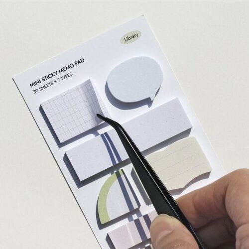 A hand using tweezers to peel a sticky note from a set of seven diverse, pastel-colored designs arranged on a white display sheet.
