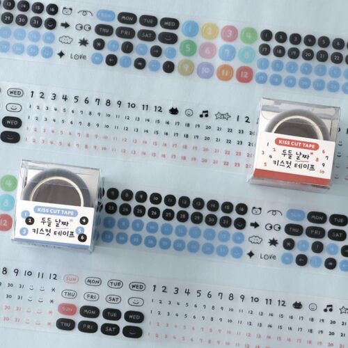 Two small, clear plastic boxes with black tape rolls inside sit atop a colorful, stickered planner page filled with doodle-themed icons and dates.