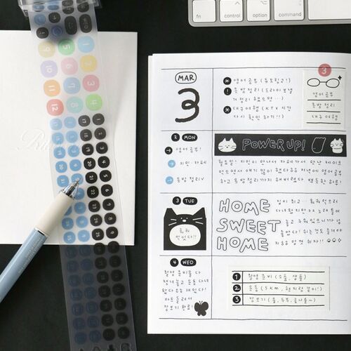 This image showcases a detailed view of a handwritten planner page with doodle date stickers, alongside a transparent sheet of colorful sticker icons and a white pen, all set against a dark surface.