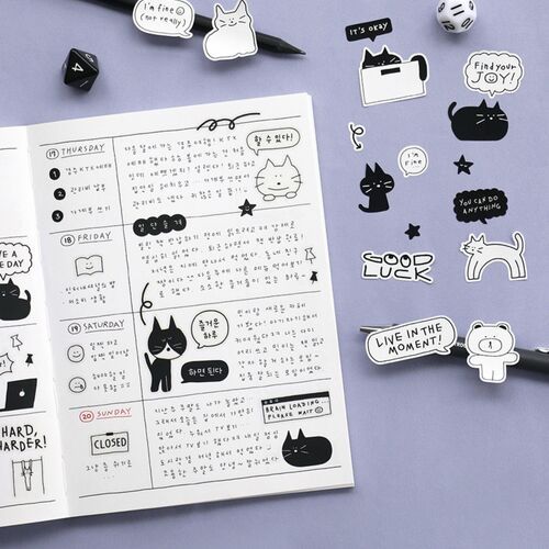 Black and white doodle stickers featuring cute cats, stars, and motivational phrases are displayed on a pastel purple surface, alongside an open journal.