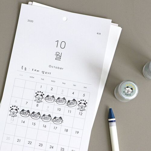 The image showcases a white calendar page with playful cat illustrations marking dates, accompanied by small stamp containers and a pen on a neutral background.