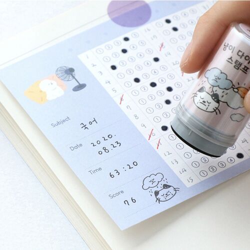 The stamp features a cute, illustrated cat design with playful expressions, positioned above a journal page with a hand holding the stamp ready to use.