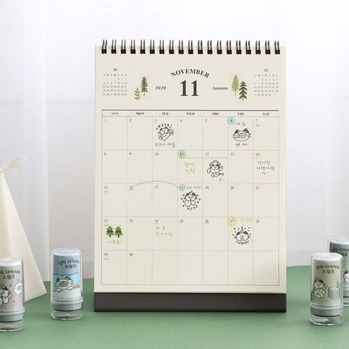 The image showcases a neatly arranged calendar with playful cat illustrations, complemented by five small, cute stamp containers on a green surface.