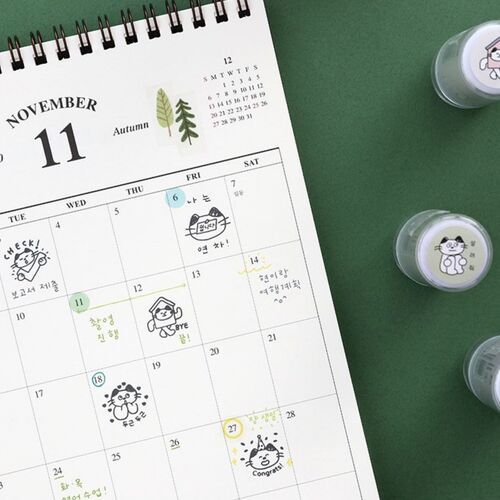 Three small, round self-inking stamps featuring cute cat illustrations are arranged on a green surface next to a planner with playful, handwritten notes and drawings.