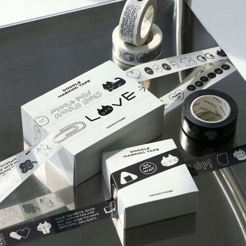 The image showcases white masking tape rolls with playful black doodle designs, arranged on a reflective metallic surface with bright, natural lighting.