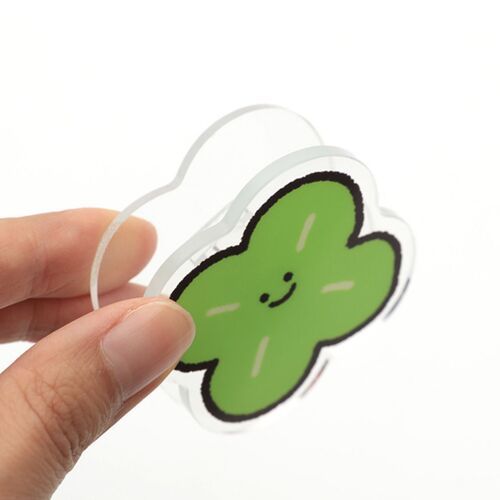 A hand holds a cheerful green clover-shaped acrylic magnet clip with a cute smiling face, set against a clean white background.