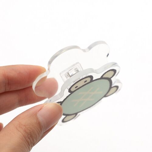 A playful acrylic magnet clip featuring a cute turtle design, held between fingers against a bright white background, highlighting its glossy finish.