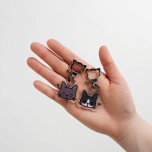 Two playful tuxedo cat emoji acrylic key rings with adorable facial expressions and cat-shaped metal clips are held against a plain white background.