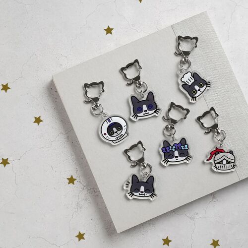 A close-up of five tuxedo cat emoji acrylic key rings with vibrant colors and playful expressions, arranged on a white textured surface.