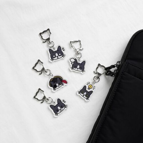 Four tuxedo cat emoji acrylic key rings with playful expressions spill out from a black bag onto a white surface, showcasing vibrant colors and detailed faces.