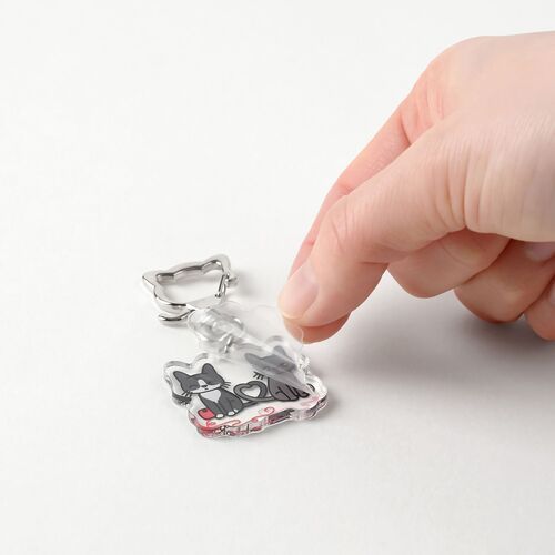 The image shows a hand peeling off a protective film from a colorful acrylic tuxedo cat emoji key ring with a playful design.