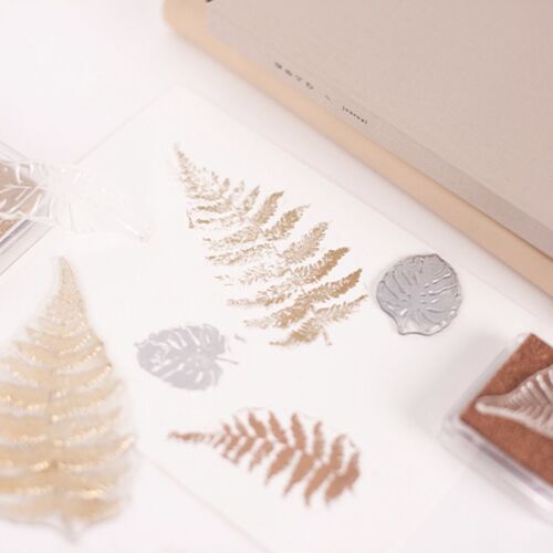 Transparent silicone stamps with intricate leaf and fern patterns are displayed alongside a neutral-toned ink pad on a white surface, highlighting their detailed design and versatility.