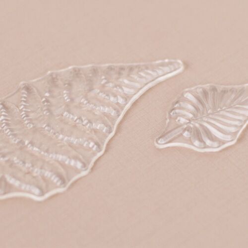 Two transparent silicone stamps with intricate leaf and fern patterns rest on a soft pink background, highlighting their flexible, detailed design.