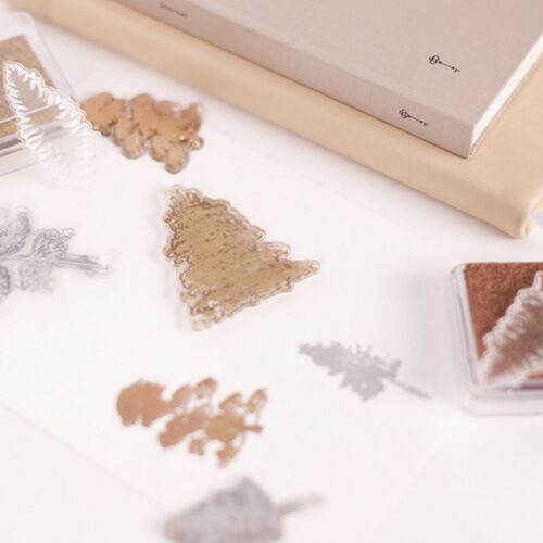 Clear silicone stamps with detailed leaf and tree designs are arranged on a white surface, accompanied by ink pads and packaging elements.