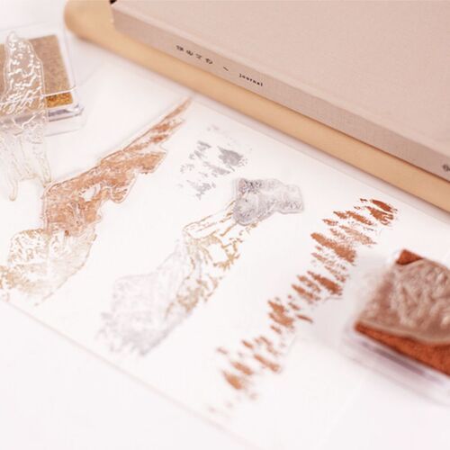 A soft pink-toned workspace showcasing stamped leaf and mountain designs on paper, highlighting the stamps' detailed and versatile nature.