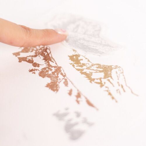 A close-up view of a hand pressing a copper-colored stamped mountain scene onto white paper, emphasizing detailed outdoor imagery.