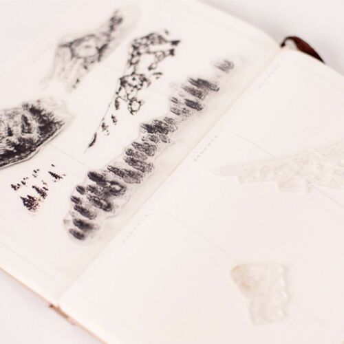 A close-up of the clear silicone stamps on an open notebook showcases detailed black ink impressions of mountain, tree, and leaf designs.