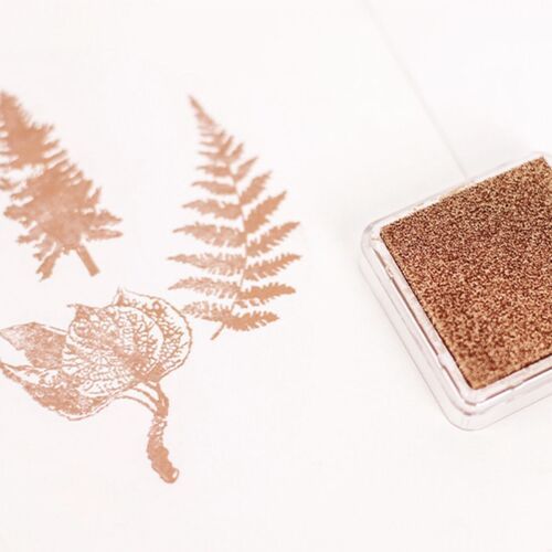 A metallic bronze shimmer stamp pad with a textured surface, positioned beside stamped leaf and fern images on white paper, highlighting its elegant finish.