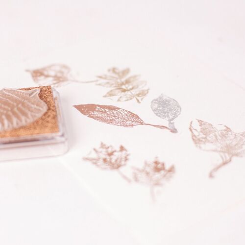 A bronze metallic shimmer stamp pad with a clear plastic lid, positioned on a white surface alongside stamped leaf patterns in matching tones.