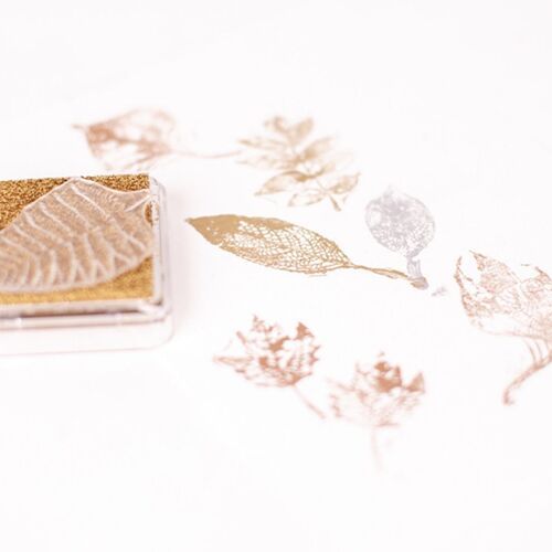 A metallic shimmer stamp pad in a soft gold hue, positioned on a white surface with delicate stamped leaf patterns in matching metallic tones.