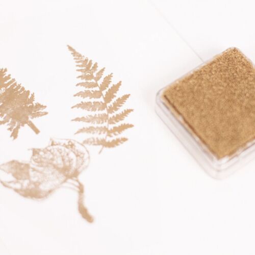 The metallic shimmer stamp pad in bronze rests beside stamped fern and leaf images, highlighting its shiny, reflective finish and elegant design.