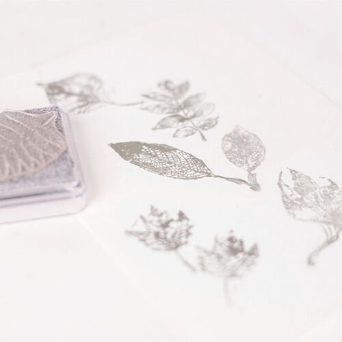 The metallic shimmer stamp pad in silver is shown with stamped leaf images on white paper, highlighting its reflective finish and detailed ink transfer.