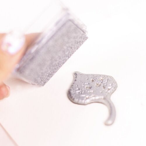 A metallic shimmer stamp pad in soft silver, with a textured surface and a small, shiny stamped image nearby, set against a clean, white background.
