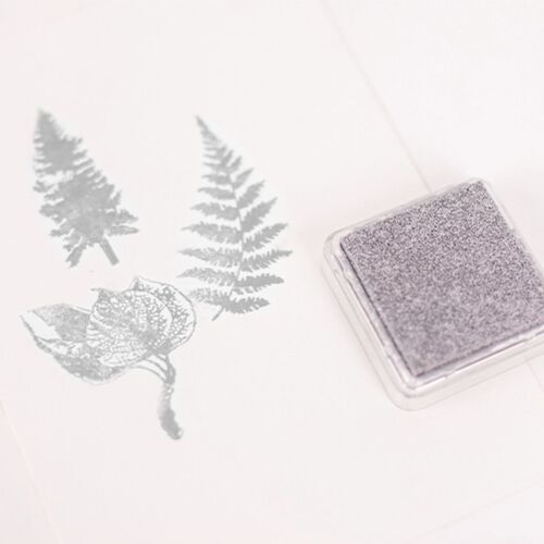 The metallic shimmer stamp pad in silver rests on a white surface, casting a subtle shadow and showcasing its textured, glittery finish.